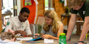 A group of young people work together at a table indoors, focusing on papers and art supplies.