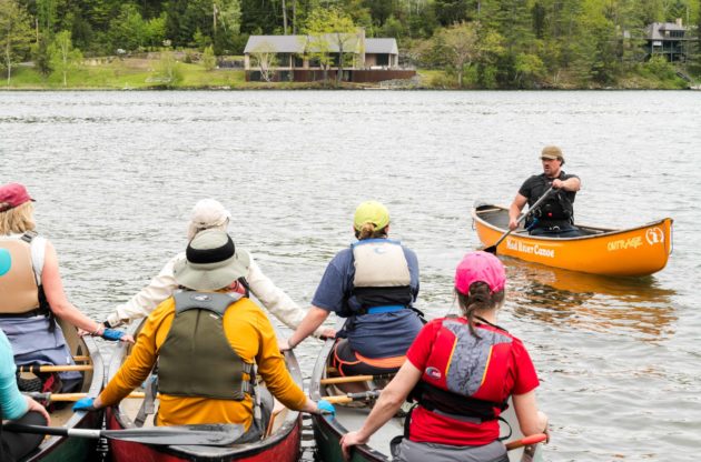 Groups of people on canoes as an instructor demonstrates.