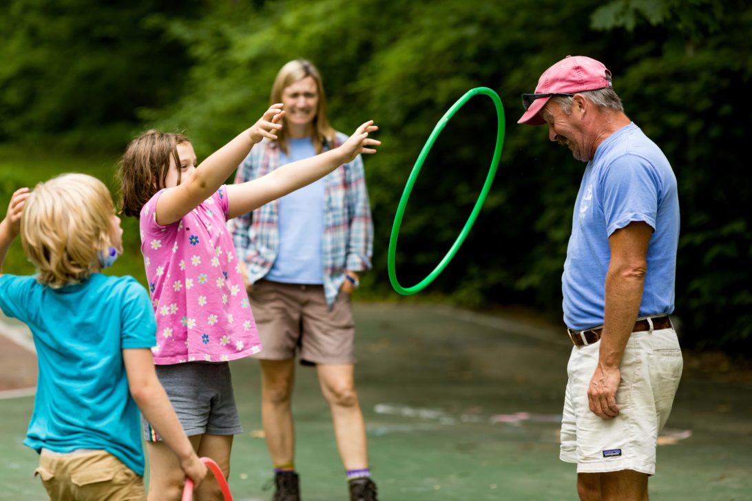 Children play with hula hoops outdoors as two adults watch. The girl tosses a green hula hoop toward an older man wearing a red cap.