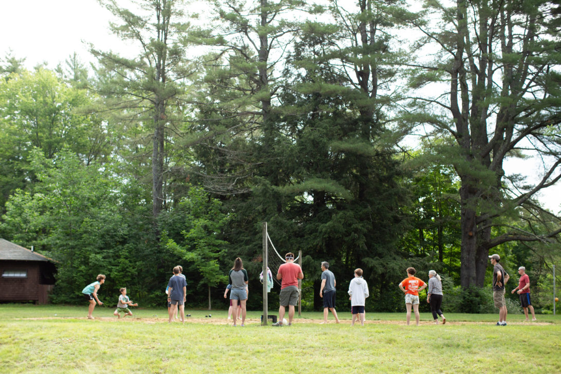 A group of people, including adults and children, are playing volleyball on a grassy area surrounded by tall trees.