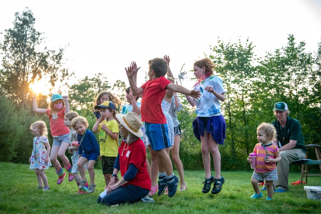 A group of children, accompanied by an adult, are playing and jumping outside on a grassy field with trees and a setting sun in the background.