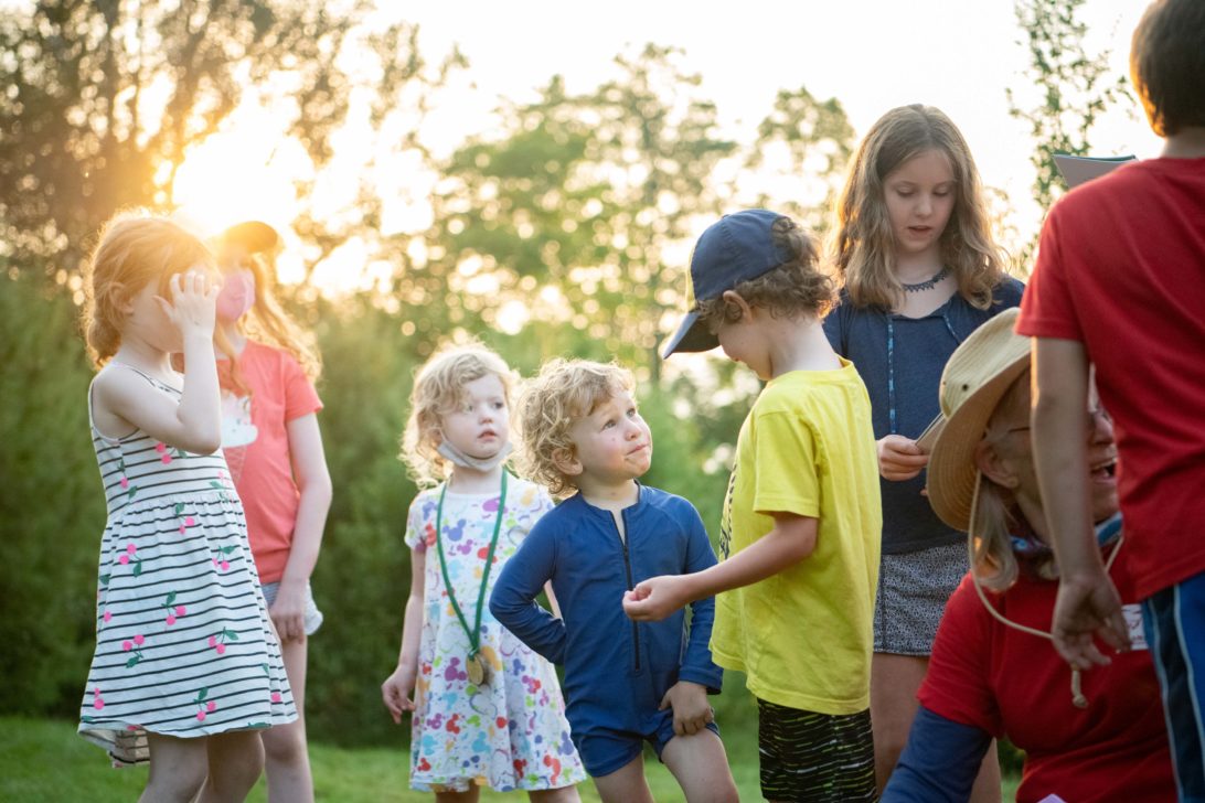 A group of children, some wearing hats, stand together outdoors in a grassy area with sunlight filtering through trees in the background.