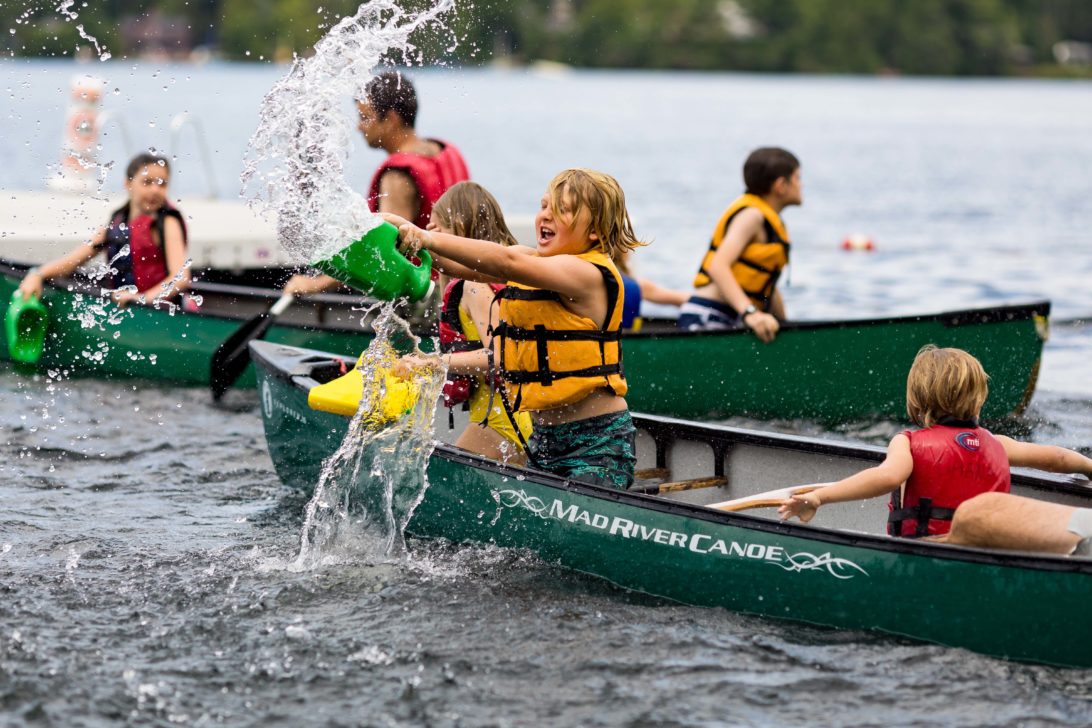Children in life jackets engage in a joyful water fight from canoes on a lake, splashing water at each other on a bright day.