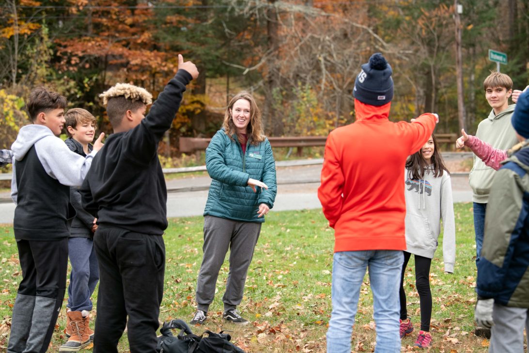 A group of children stands in a circle outdoors during autumn, smiling and gesturing with their hands, led by an adult woman wearing a teal jacket.