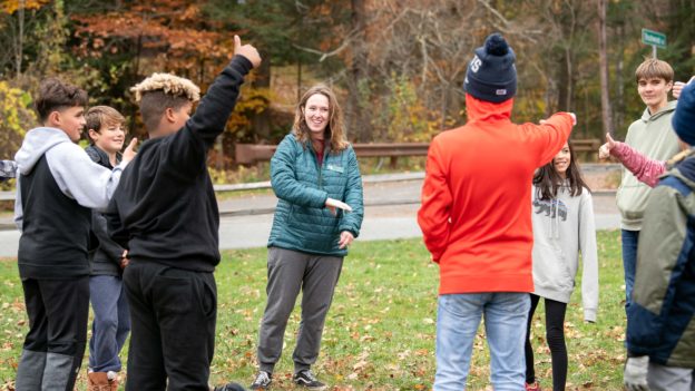 A group of children stands in a circle outdoors during autumn, smiling and gesturing with their hands, led by an adult woman wearing a teal jacket.