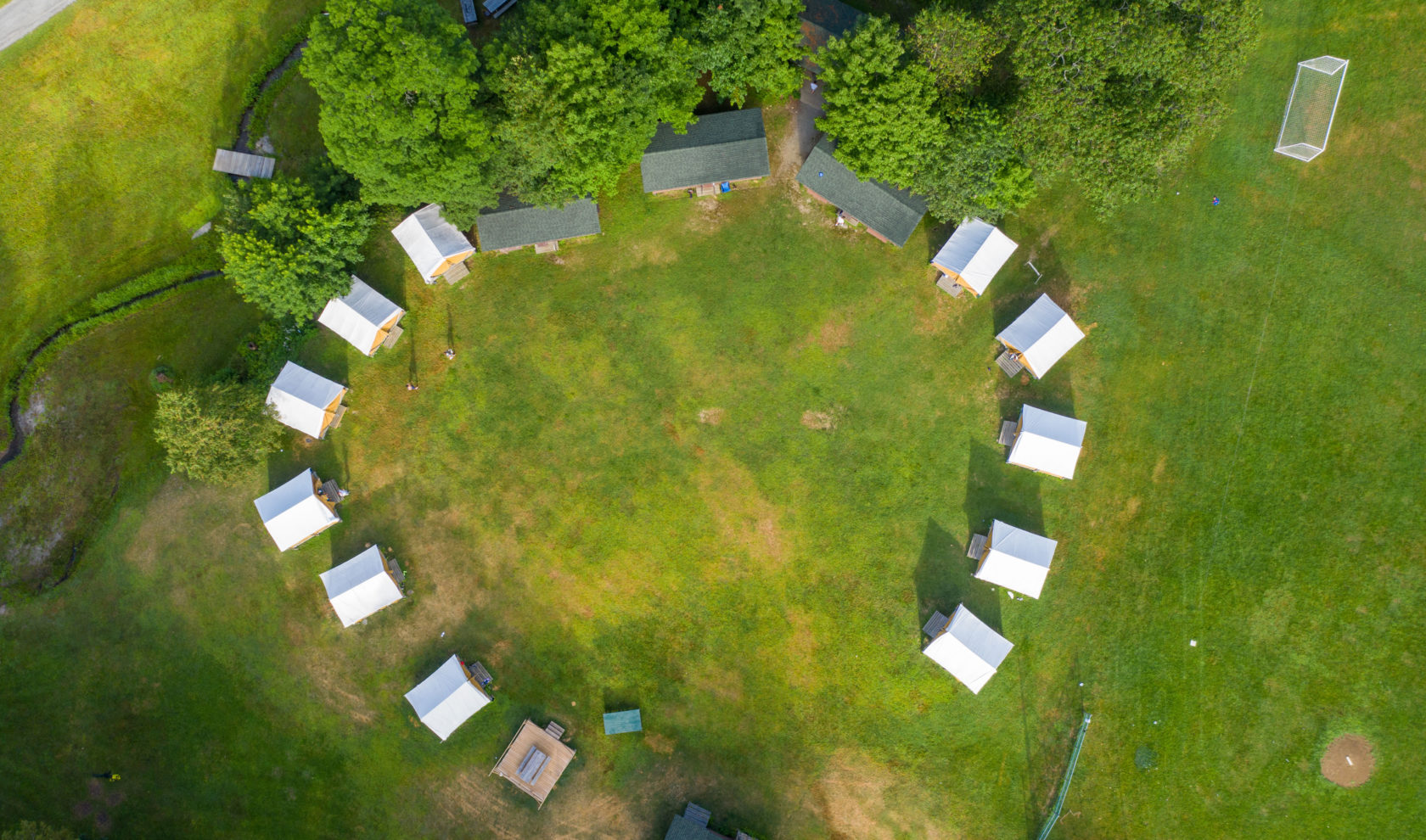 Aerial view of a circular arrangement of white tents and small cabins in a grassy clearing, surrounded by trees.