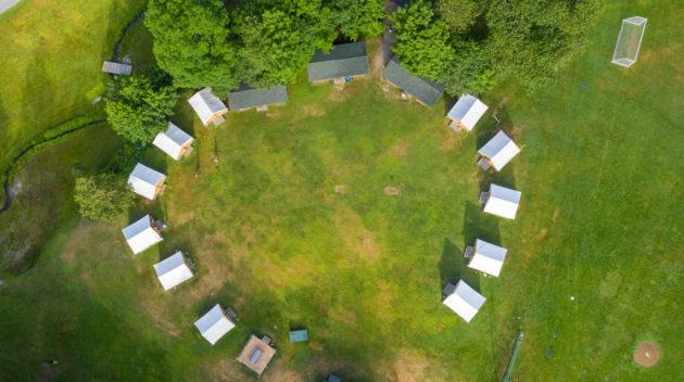 Aerial view of a circular arrangement of white tents and small cabins in a grassy clearing, surrounded by trees.