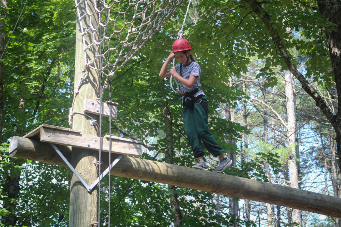 Person in safety gear balancing on a wooden beam high above the ground in an outdoor ropes course with trees in the background.