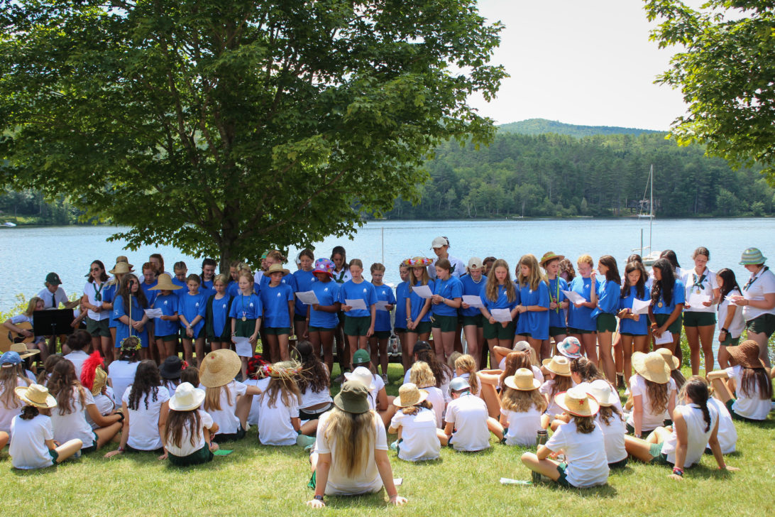 Group of children in blue and white shirts gathered outdoors near a lake, some standing under a tree and others sitting on grass, participating in an event or activity.