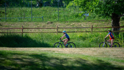 Two people riding bicycles on a path beside a wooden fence and grassy field on a sunny day.