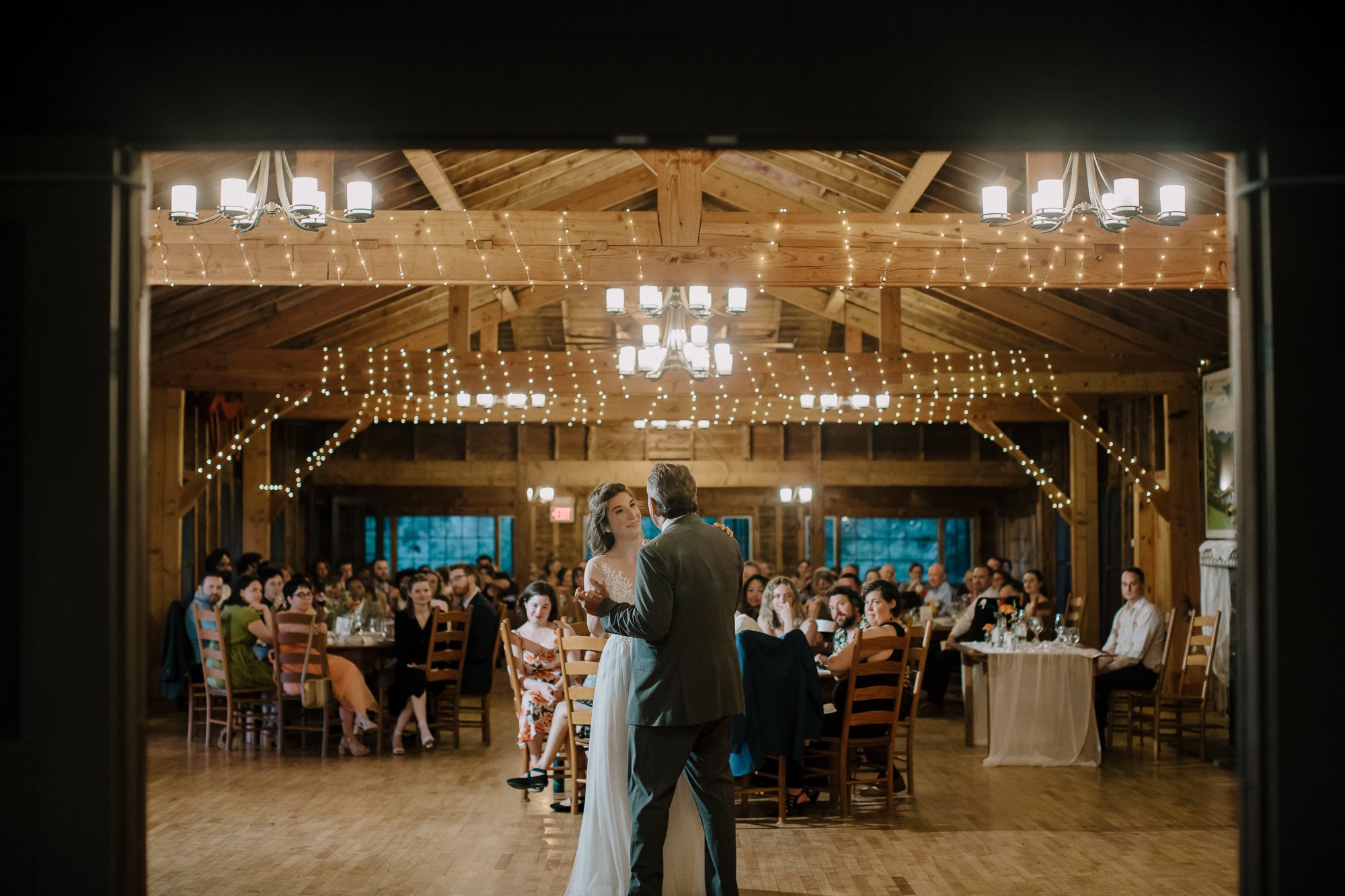 A couple dances in a rustic banquet hall with wooden beams and string lights, surrounded by seated guests who are watching attentively.