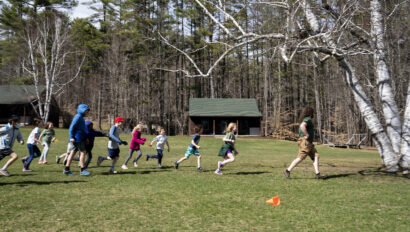 Children and an adult run outdoors on a grassy field near trees and small cabins, under clear skies.
