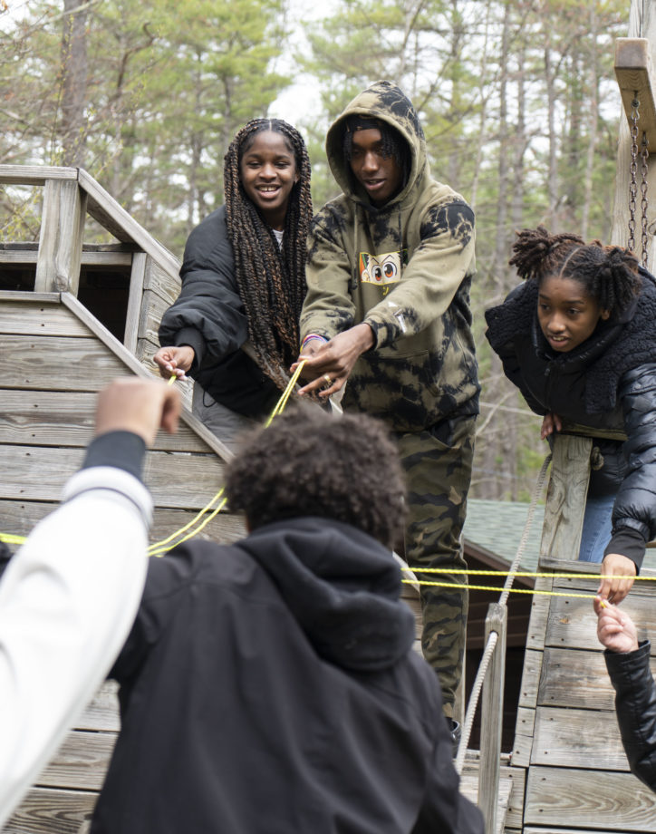 A group of people, dressed in casual outdoor clothing, are working together with ropes in a team-building exercise on a wooden structure with trees in the background.