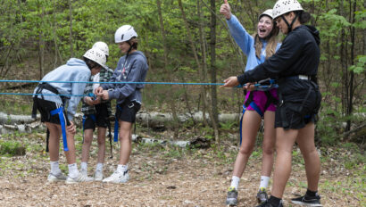 Four outdoor adventurers wearing helmets and harnesses prepare for a zipline challenge; one gives a thumbs up while the others make adjustments.