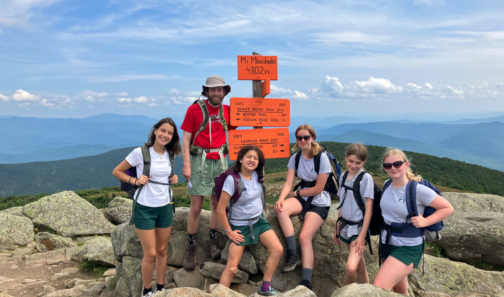 A group of hikers standing in front of a Mt. Moosilauke summit sign, indicating an elevation of 4,802 feet, with mountainous terrain in the background.