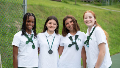 Four smiling students wearing white uniforms and green ties stand together outside near at the tennis courts, with a grassy area in the background.
