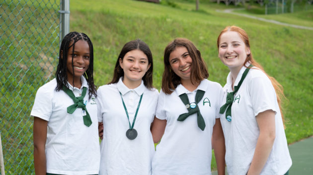 Four smiling students wearing white uniforms and green ties stand together outside near at the tennis courts, with a grassy area in the background.
