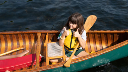 A child wearing a yellow life jacket gives two thumbs up while sitting in a wooden canoe on a lake.
