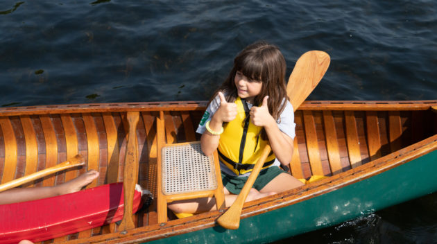 A child wearing a yellow life jacket gives two thumbs up while sitting in a wooden canoe on a lake.