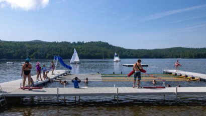 People enjoying various activities at a lakeside dock area with a slide. Sailboats are visible in the background, and the surrounding area is lush with greenery and hills.
