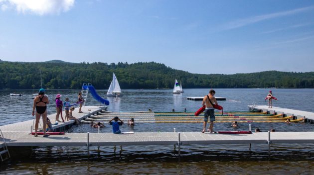People enjoying various activities at a lakeside dock area with a slide. Sailboats are visible in the background, and the surrounding area is lush with greenery and hills.