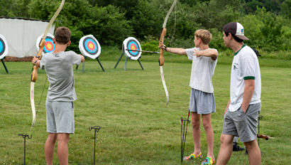 Three young people practice archery on a grassy field, aiming at colorful targets with bows. One person stands observing while two others are in mid-aim.
