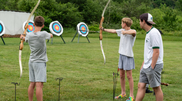 Three young people practice archery on a grassy field, aiming at colorful targets with bows. One person stands observing while two others are in mid-aim.