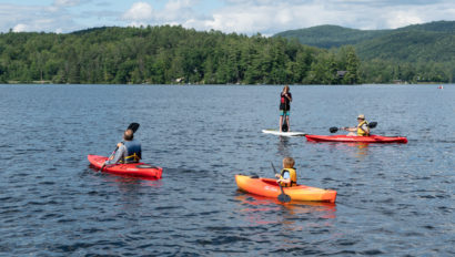 Four people are on a lake, three in kayaks, and one on a paddleboard. Forested hills and a partly cloudy sky are in the background.