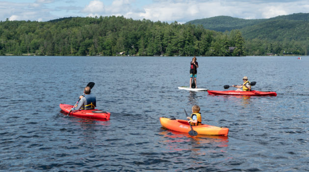 Four people are on a lake, three in kayaks, and one on a paddleboard. Forested hills and a partly cloudy sky are in the background.
