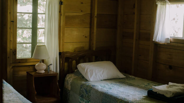 A small wooden room with two single beds, each with a quilt and pillow, separated by a nightstand with a lamp.