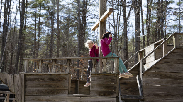 Two children sit on a wooden play structure shaped like a ship in a forest setting. Both are holding onto a post, looking outward.
