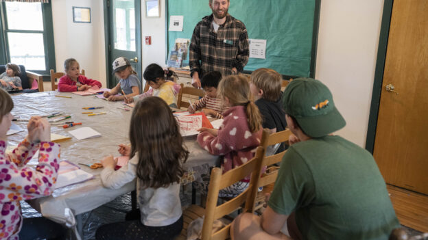 A group of children sits at a table drawing and coloring, while an adult stands at the head of the table smiling and observing them.