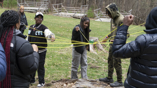 A group of people outdoors participating in a team-building activity that involves holding and weaving yellow strings.