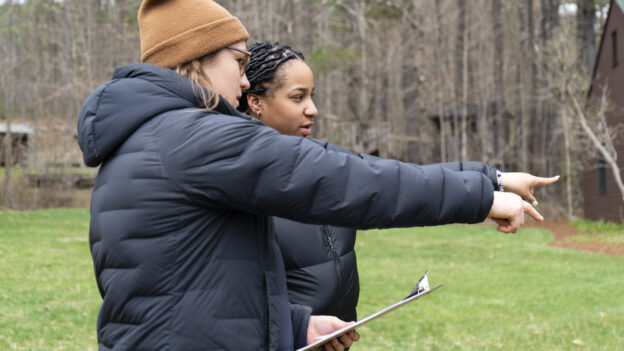 Two people wearing winter jackets and beanies stand outdoors, one holding a clipboard, both pointing towards the same direction.