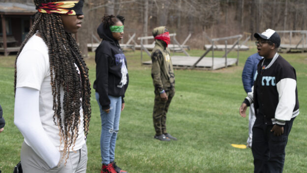 Several people stand blindfolded outdoors on a grassy area. One person appears to be guiding the others, who are spaced out and facing different directions.