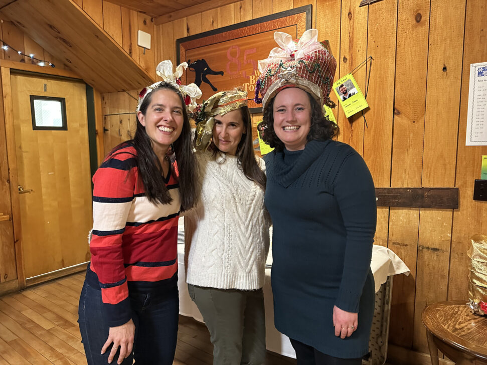 Three people standing indoors wearing gift-wrapped hats, smiling at the camera. The room has wooden walls and a festive atmosphere.