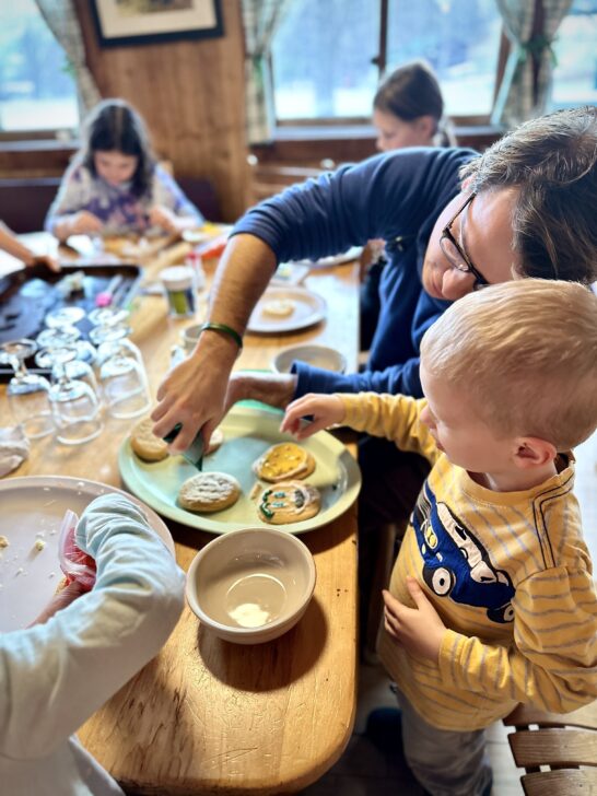 An adult and child are decorating cookies at a table with other children engaged in the same activity. Various cookie decorating supplies are scattered on the table.