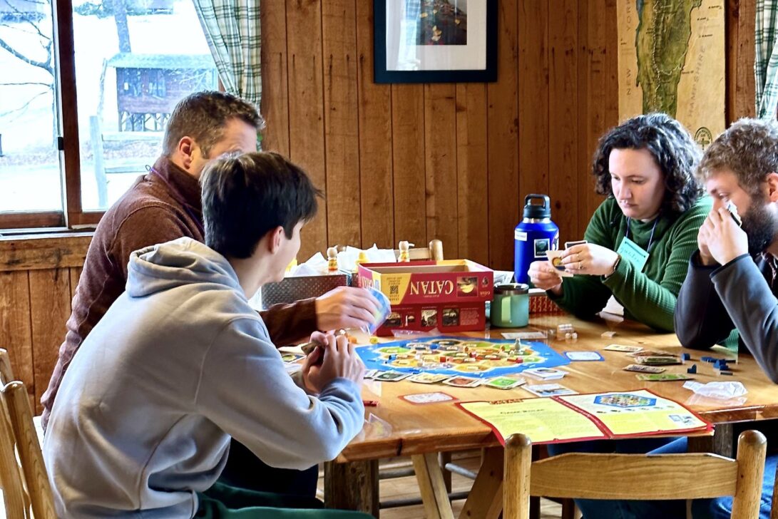 Four individuals are seated around a wooden table, intently playing a board game with various cards and pieces. The setting appears to be a cozy, rustic room with wood paneled walls and large windows.