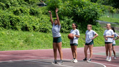 A group of young women wearing athletic attire stands on an outdoor basketball court. One woman is shooting a basketball while the others wait in line behind her.
