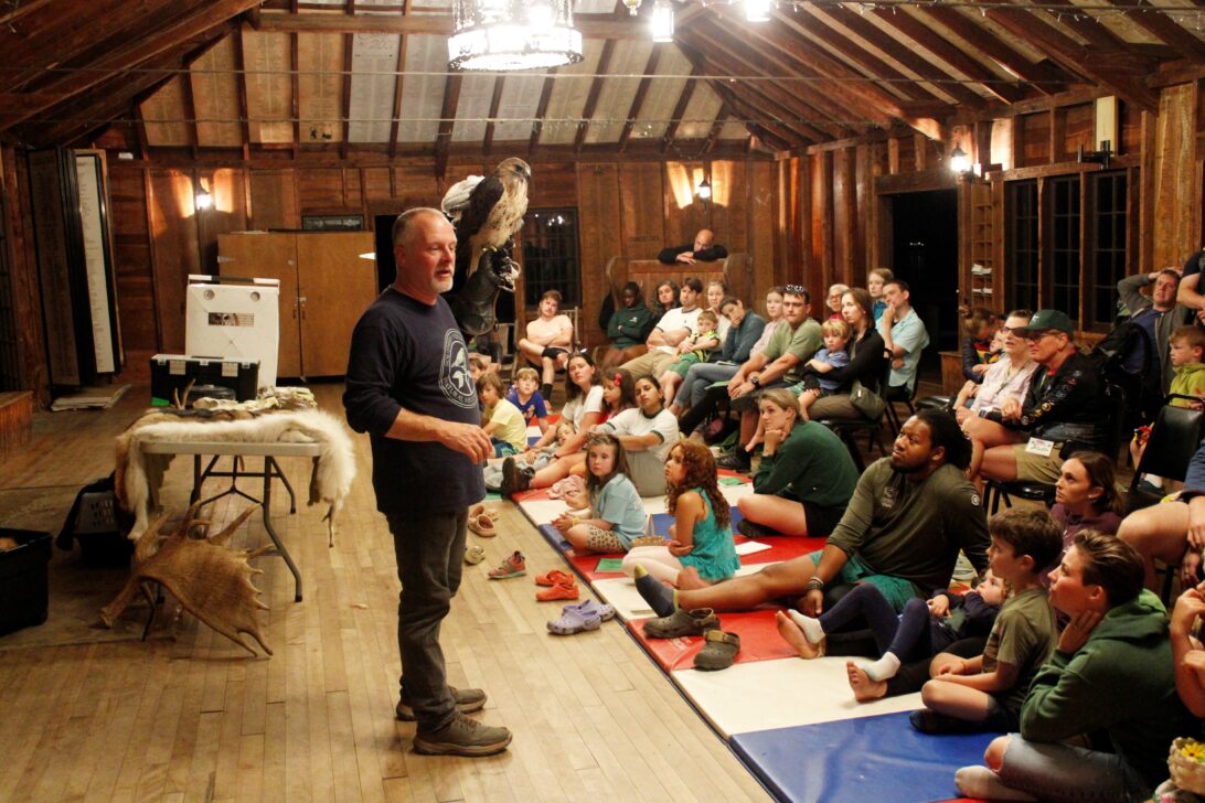 A man stands speaking to an attentive audience in a rustic, wooden room, with various teaching materials and a bird on display behind him.