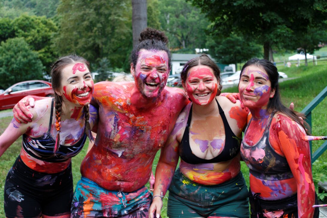 Four people, each covered in colorful paint, stand arm-in-arm with smiles outdoors in a grassy area with trees in the background.