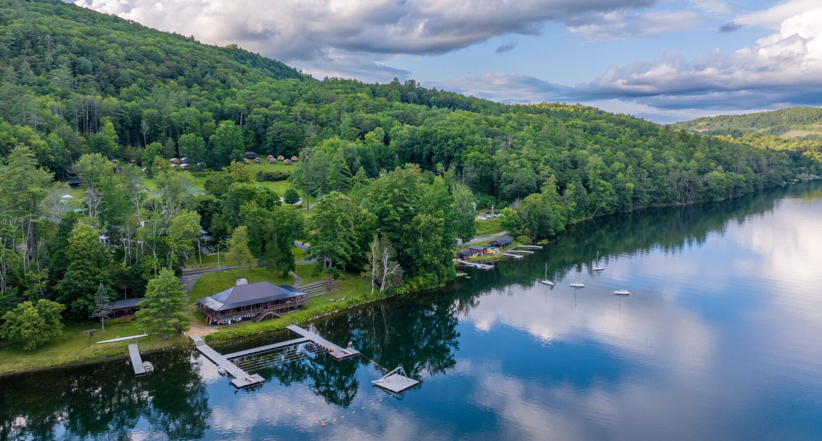 A serene lakeside scene with docks extending into calm water, surrounded by lush green trees and forested hills under a partly cloudy sky. A large building is visible near the shoreline.