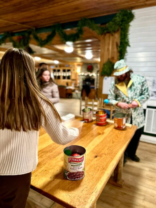 Three people engaged in a maple syrup demonstration, pouring syrup into cans labeled "Pure Maple Syrup" on a wooden table, with a rustic background and greenery decorations.