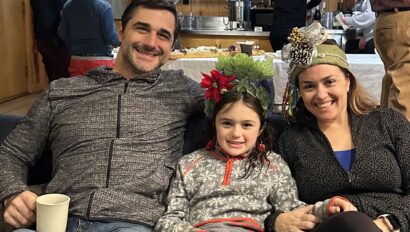 A man, young girl, and woman sit closely on a couch, smiling at the camera. The child and woman are wearing festive headbands.