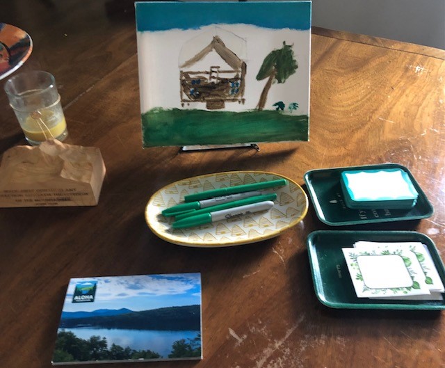 A wooden table with assorted items: a painting, a half-drunk beverage, a small plaque, a postcard, green pens in a dish, and trays holding notepads and stationery.