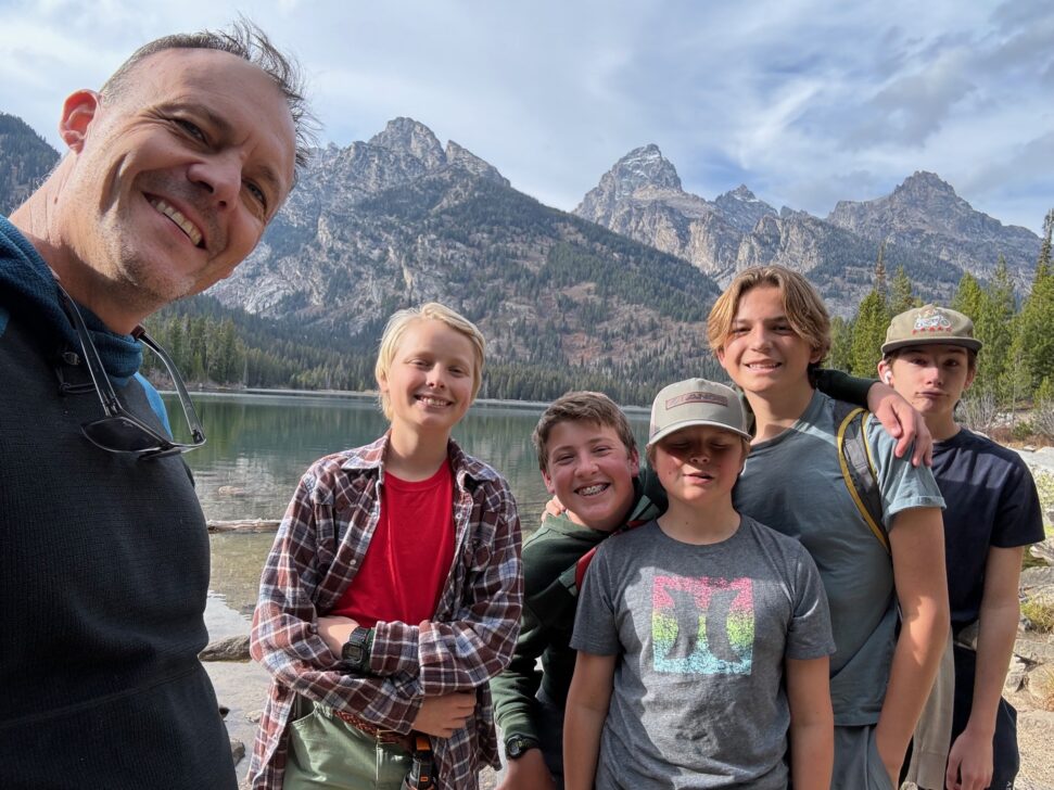 A group of six people, including five young individuals and one adult, stand and smile by a lake with mountains and trees in the background.