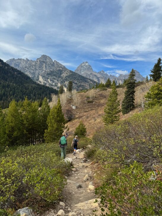 Two hikers walk along a rocky path surrounded by greenery, with mountains and a partly cloudy sky in the background.