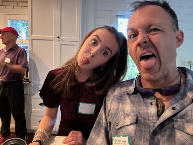 Two people are making funny faces at the camera at an indoor gathering, while another person in a red cap stands in the background.