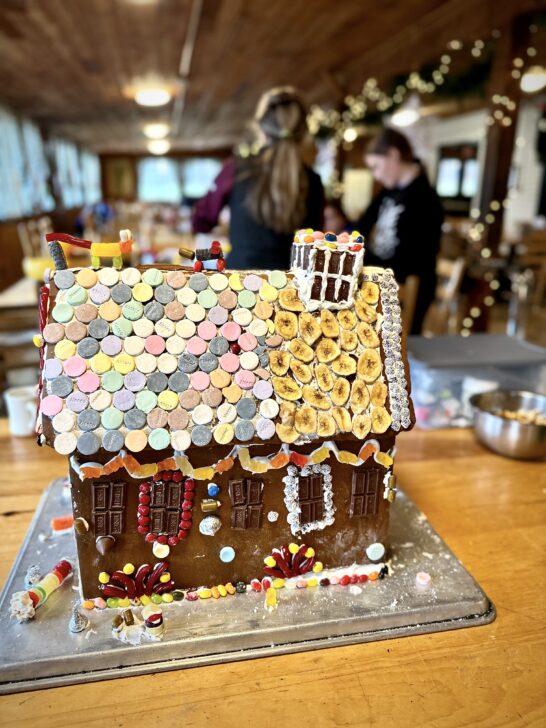 A gingerbread house decorated with colorful candies and icing on a wooden table. In the background, people are slightly blurred, working in a room with festive lights.