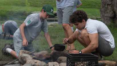 Two boys crouch by a campfire. One stirs a pot on the fire while the other observes.